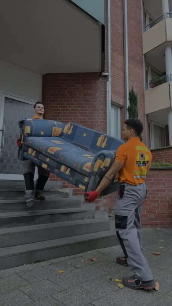 Professionelle Haushaltsauflösung in Aachen: Zwei Mitarbeiter des Teams Jung und Fix tragen ein großes, gemustertes Sofa vorsichtig die Außentreppe eines Wohnhauses hinunter.