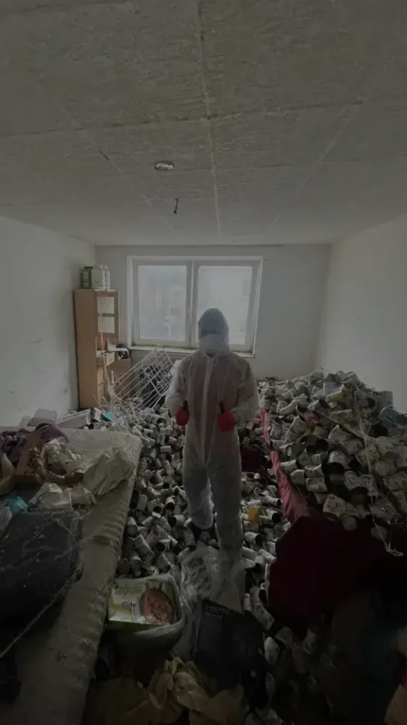 Professionelle Messie Entrümpelung in Aachen: Ein Mitarbeiter von Jung und Fix im weißen Schutzanzug steht inmitten eines stark vermüllten Zimmers voller Abfall und Dosen.