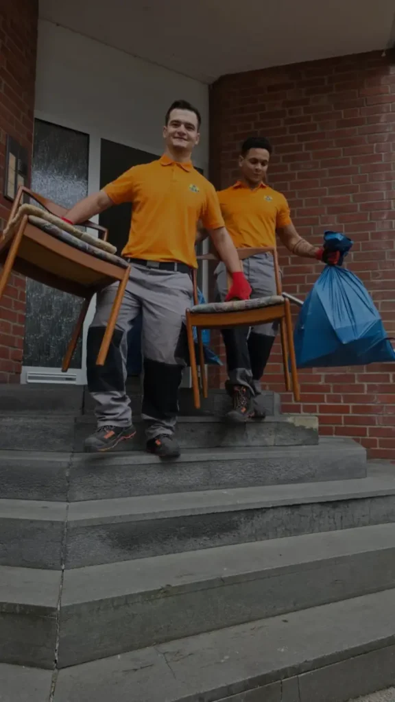 Zwei freundliche Mitarbeiter von Jung und Fix tragen bei einer professionellen Entrümpelung in Aachen alte Stühle und gefüllte Müllsäcke die Treppe eines Wohnhauses hinunter.